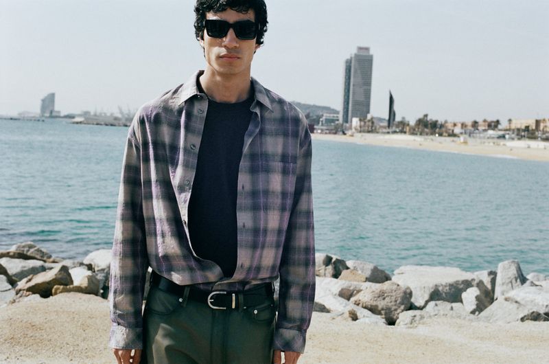 Model wearing dark sunglasses and an unbuttoned plaid shirt standing outdoors by the ocean and sandy beach