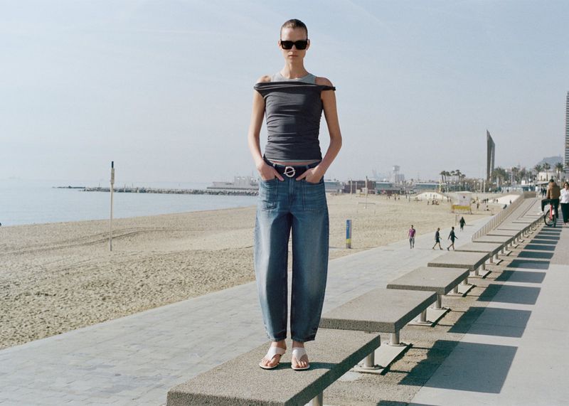 Adult woman in sunglasses wearing a gray top and denim pants standing on a concrete step with a beach and city skyline in background.