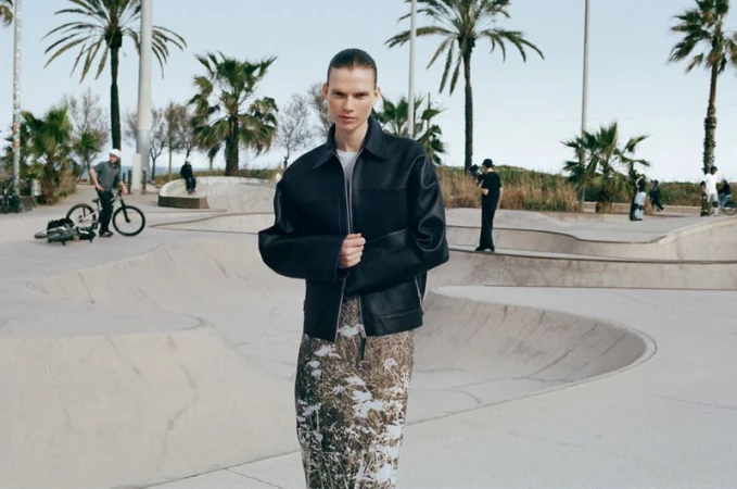 Model wearing a black jacket and patterned skirt standing outdoors near a skate park with palm trees in the background.