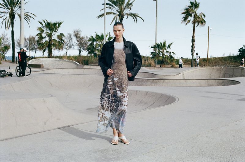 Model wearing a black leather jacket and patterned dress standing in a concrete skatepark with palm trees