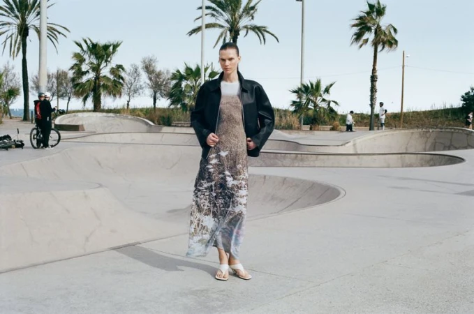 Model wearing a black leather jacket and patterned dress standing in a concrete skatepark with palm trees