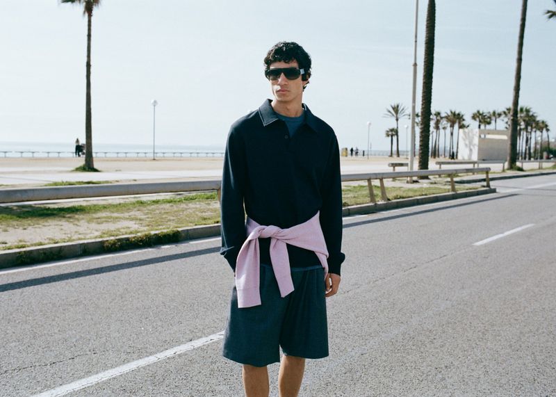 A man wearing sunglasses, a dark polo shirt, and shorts standing on a paved road with palm trees in the background.