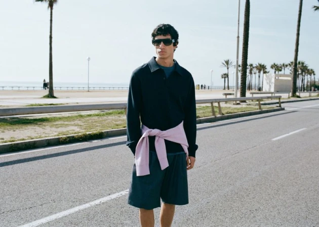 A man wearing sunglasses, a dark polo shirt, and shorts standing on a paved road with palm trees in the background.