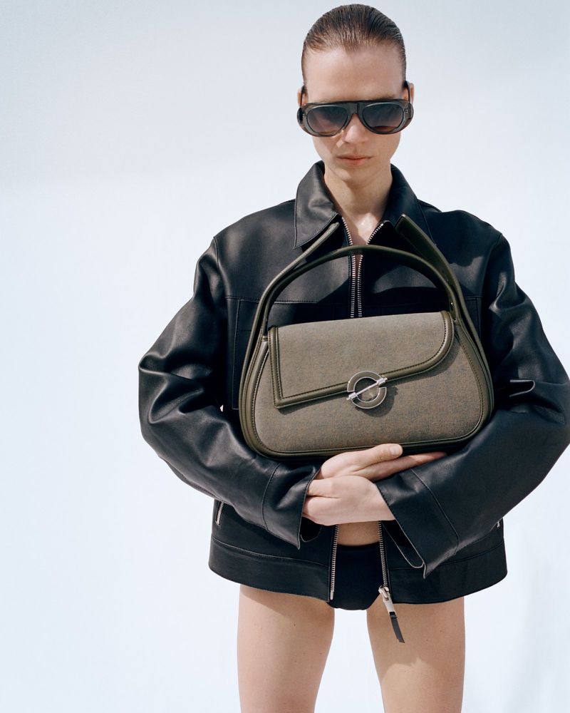 Model in dark sunglasses and black leather jacket holding an olive handbag against a plain background
