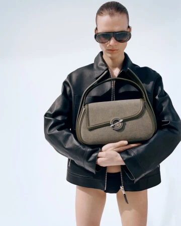Model in dark sunglasses and black leather jacket holding an olive handbag against a plain background