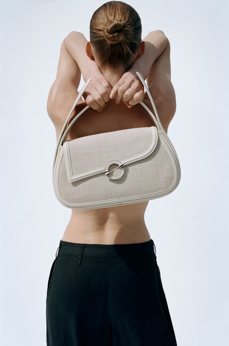 Model holding a structured light-colored handbag behind their back against a clean white backdrop.