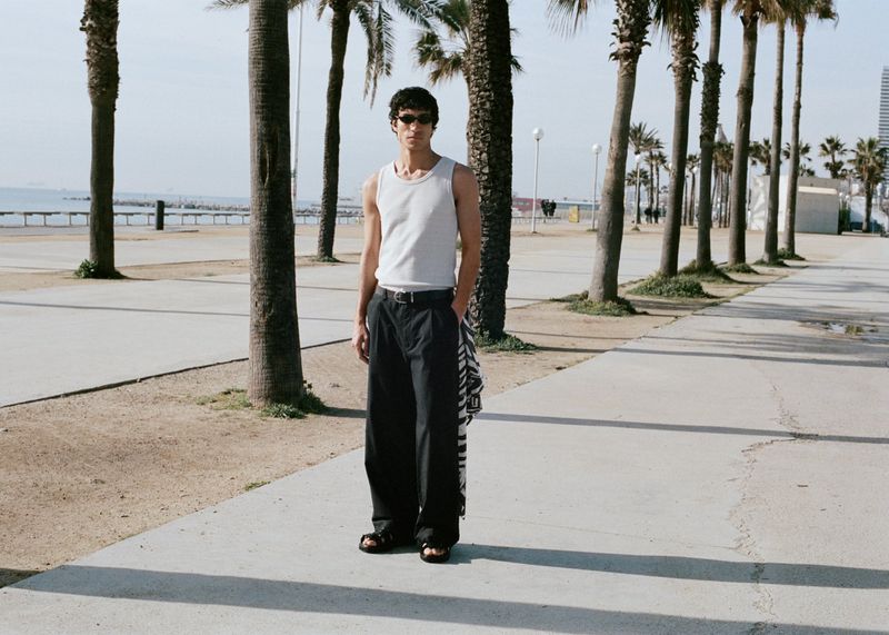Model wearing a white tank top and black trousers standing on a palm-lined promenade by the coast.