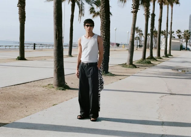 Model wearing a white tank top and black trousers standing on a palm-lined promenade by the coast.