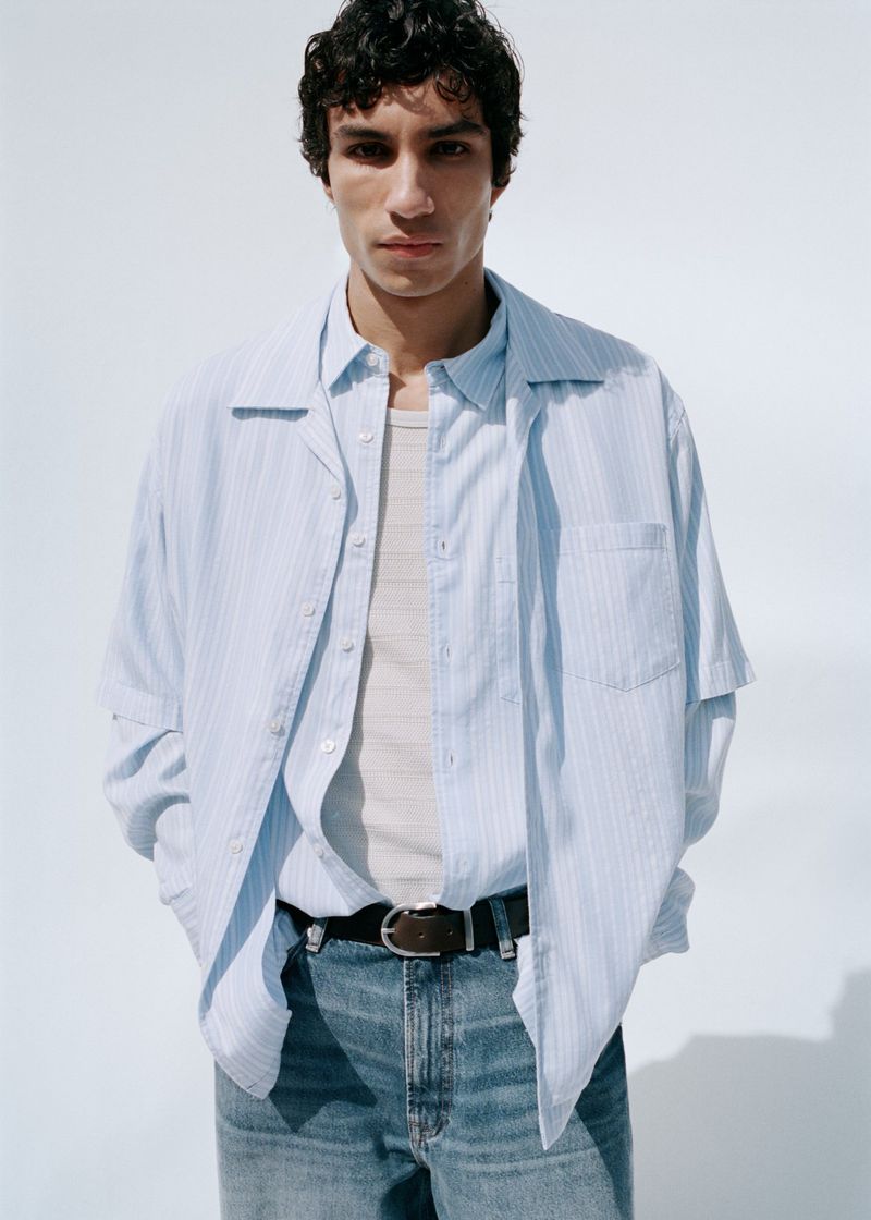Male model wearing a light blue striped shirt over a tank top and blue jeans against a white background