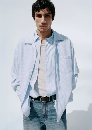 Male model wearing a light blue striped shirt over a tank top and blue jeans against a white background