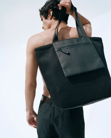 Shirtless man carrying a large black mesh tote bag over his shoulder against a plain white background.