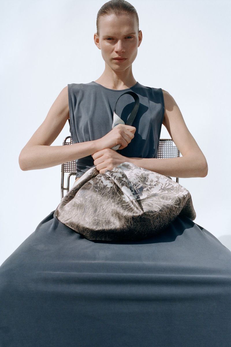 Model seated on a metal chair wearing a dark grey dress holding a patterned fabric bag against a white background