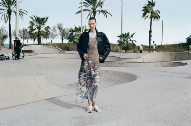 Model wearing a black leather jacket and patterned dress standing in a concrete skatepark with palm trees