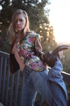 Coco Gordon Moore wearing a floral brocade jacket and blue jeans, holding her raised boot while posing by an outdoor railing.