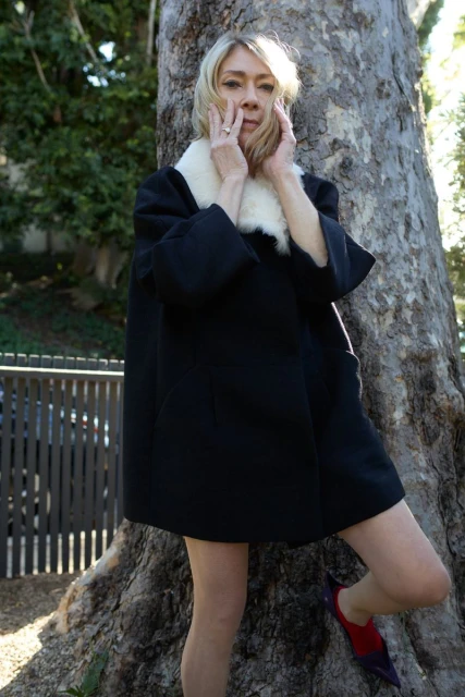 Woman with blonde hair poses in front of a large tree, wearing a black coat with a white fur collar and colorful pointed-toe shoes.