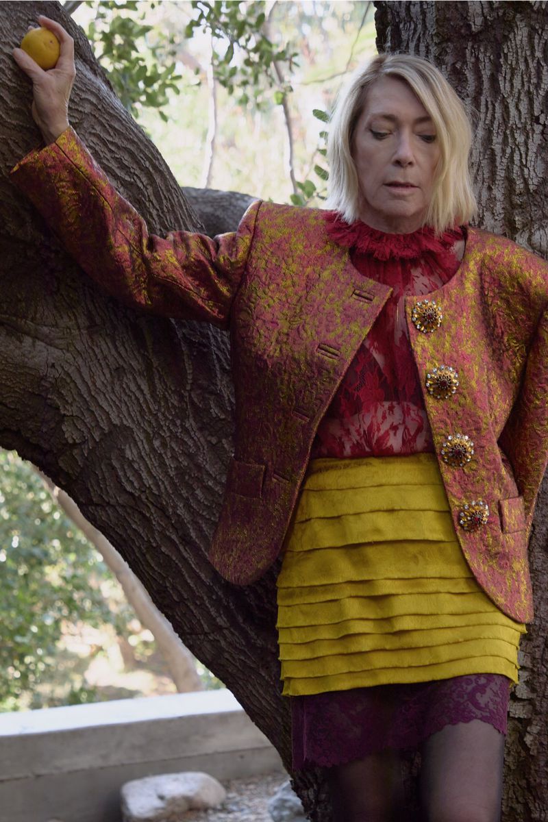 Stylish adult woman in patterned gold jacket and yellow tiered skirt poses with a lemon against a tree in an outdoor setting.