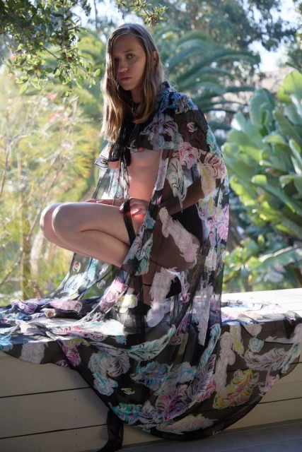 Blonde adult woman wearing a floral robe crouching on a white ledge outdoors surrounded by green foliage in a medium eye-level shot.