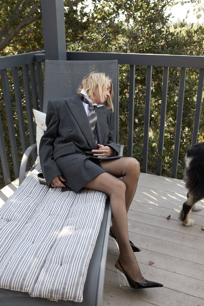 Woman in grey suit jacket, tie, and sheer tights sits on striped lounge chair on deck; dog nearby, trees in background. Fashion photography.