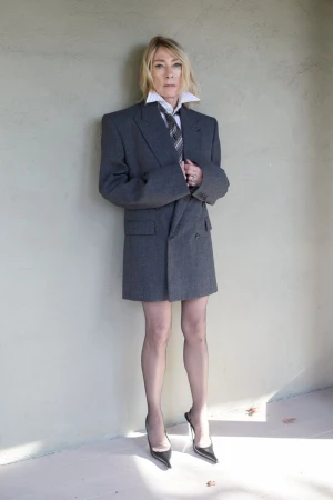 Elegant blonde woman in an oversized grey blazer, white collared shirt, and black heels posing indoors against a neutral wall.