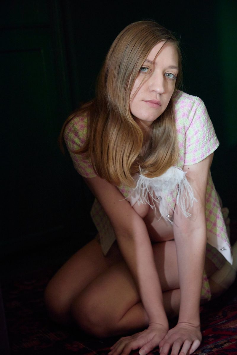 Blonde woman kneeling indoors, wearing a light pink checkered dress with feather details, against a dark backdrop with a red patterned floor.