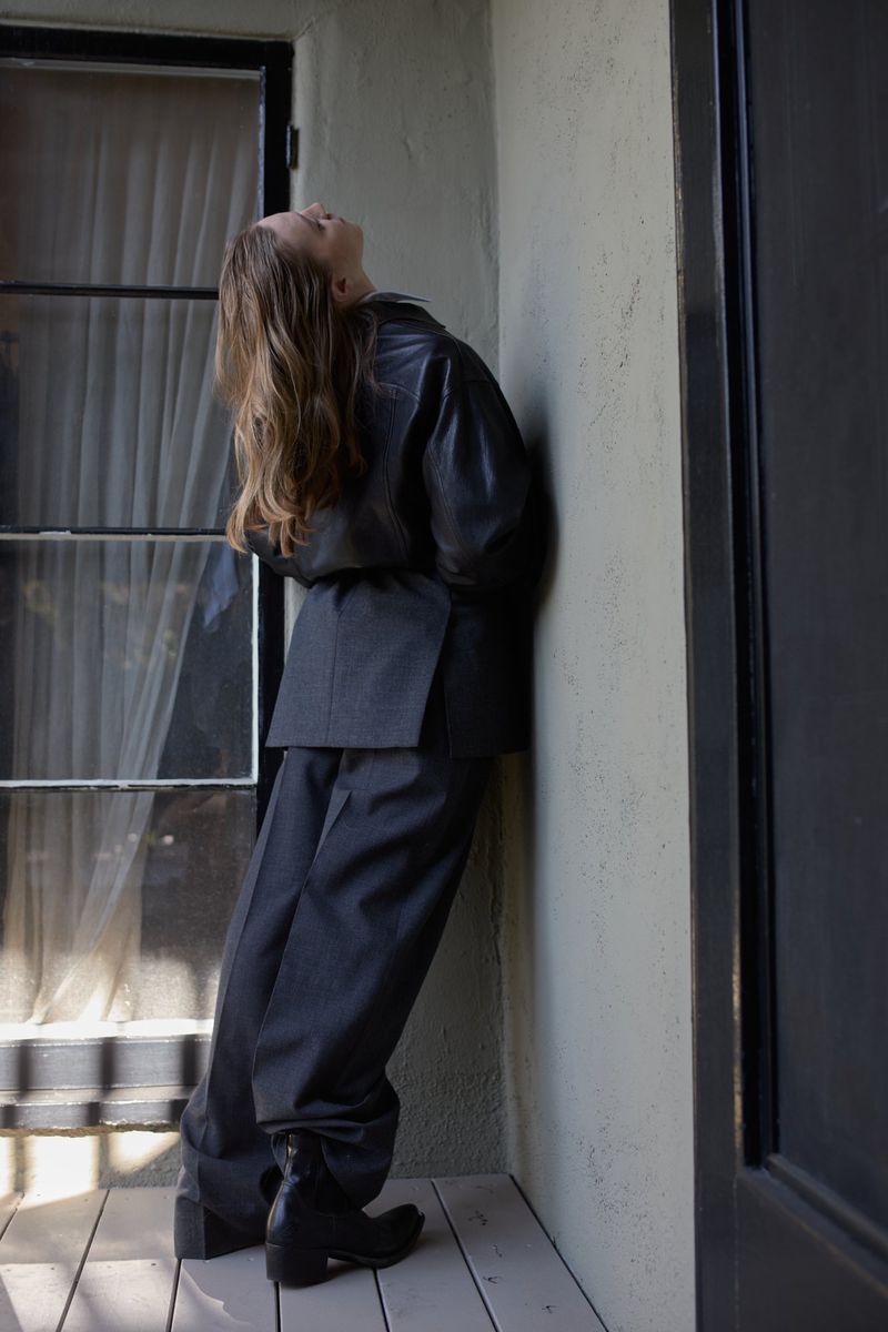 A woman with long brown hair wears an oversized suit while leaning against a wall indoors near a window with sheer curtains.