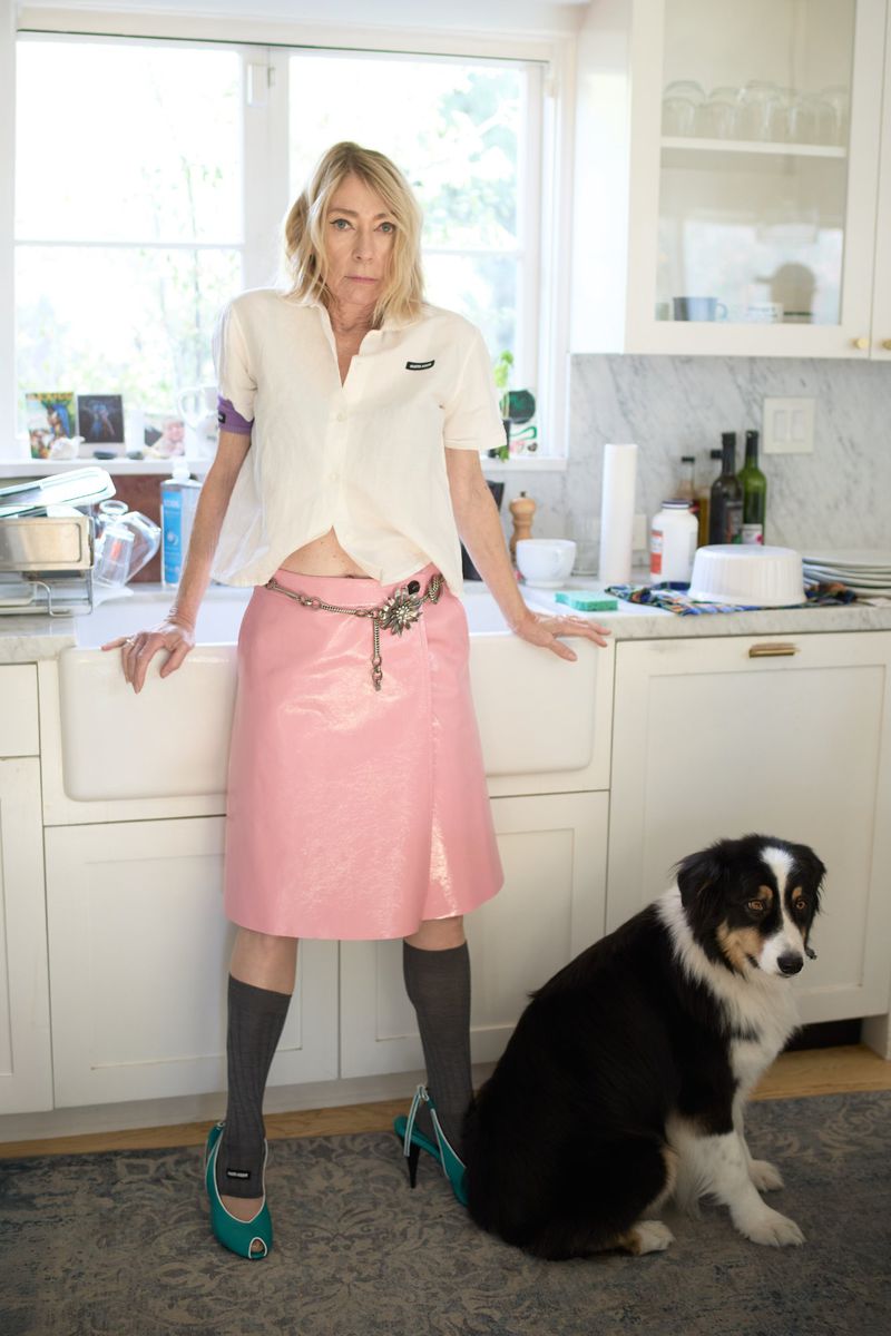 Stylish adult woman poses indoors with an Australian Shepherd dog, wearing a pink skirt, white shirt, turquoise heels, and grey socks.