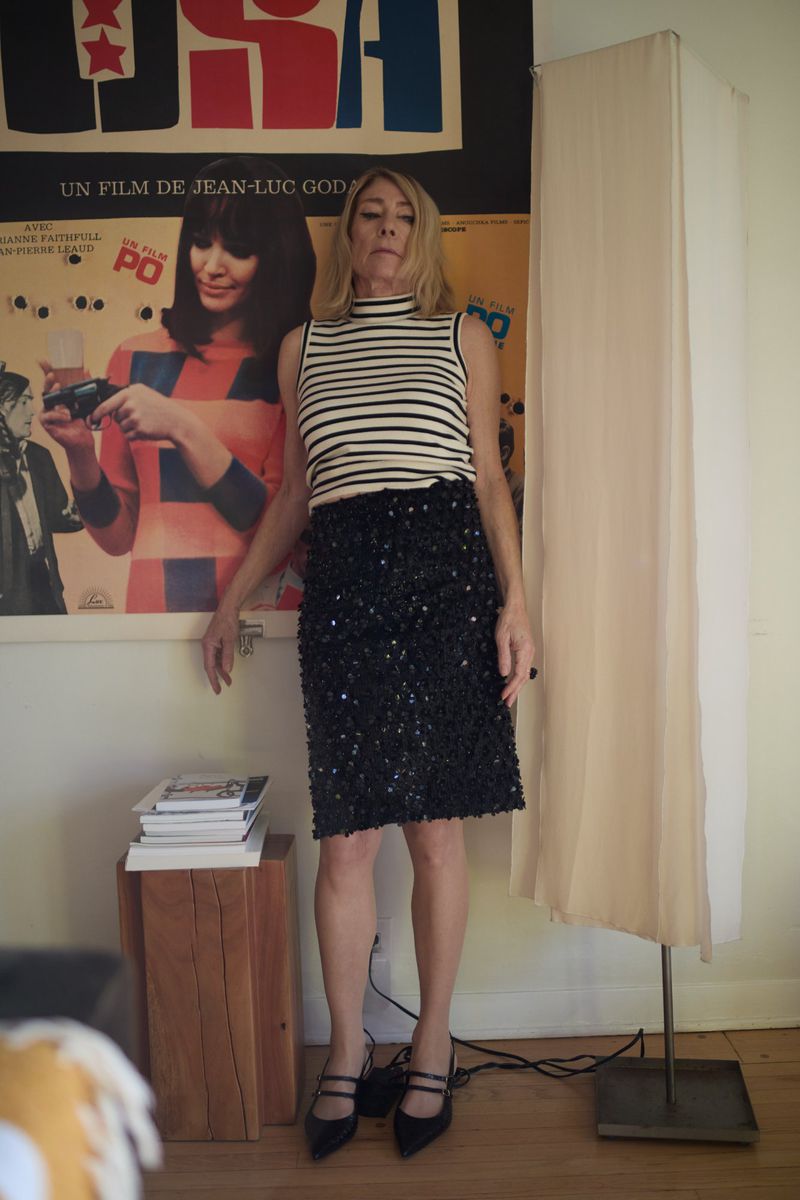 Adult woman in striped top and black skirt poses in front of a vintage film poster near a wooden side table and lamp.