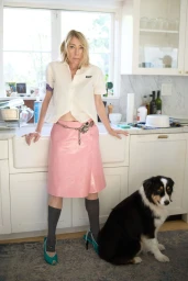 Kim Gordon wearing a white shirt and pink skirt standing by a kitchen sink next to an Australian Shepherd dog.