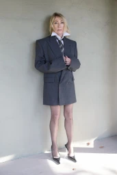 Kim Gordon wearing an oversized grey blazer, white shirt, and tie against a plain concrete wall.