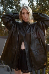 Kim Gordon wearing an oversized black leather jacket with hands behind her head on an outdoor balcony.