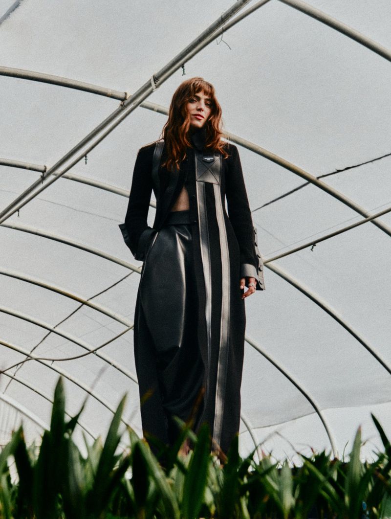 Low angle portrait of Bella Maclean wearing a dark long coat inside a greenhouse structure for Elle UK