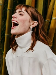 Portrait of actress Bella Maclean laughing aloud in a white turtleneck against a vertical bamboo background for Elle UK.