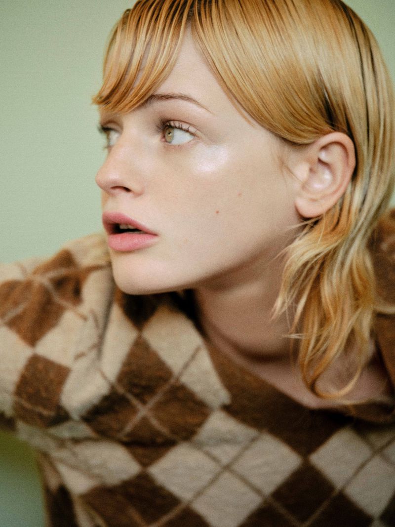 Close-up of a blonde woman with short hair wearing a brown argyle sweater, looking to the side in a soft, diffused light.