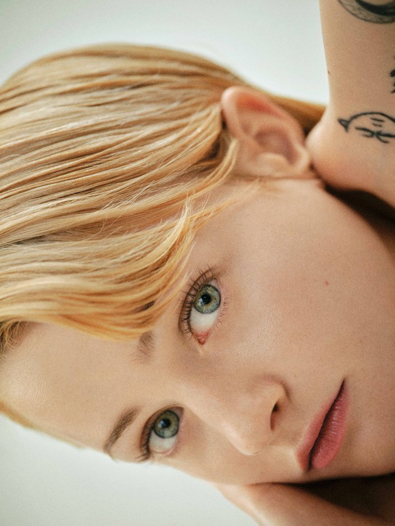 Close-up portrait of Esther McGregor with short blonde hair and arm tattoos resting her chin on her hand against a light background.