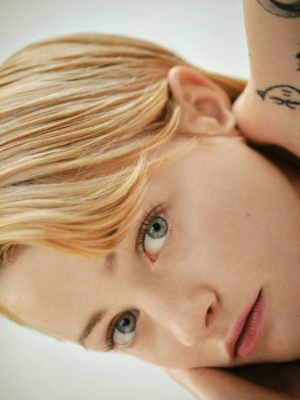 Close-up portrait of Esther McGregor with short blonde hair and arm tattoos resting her chin on her hand against a light background.