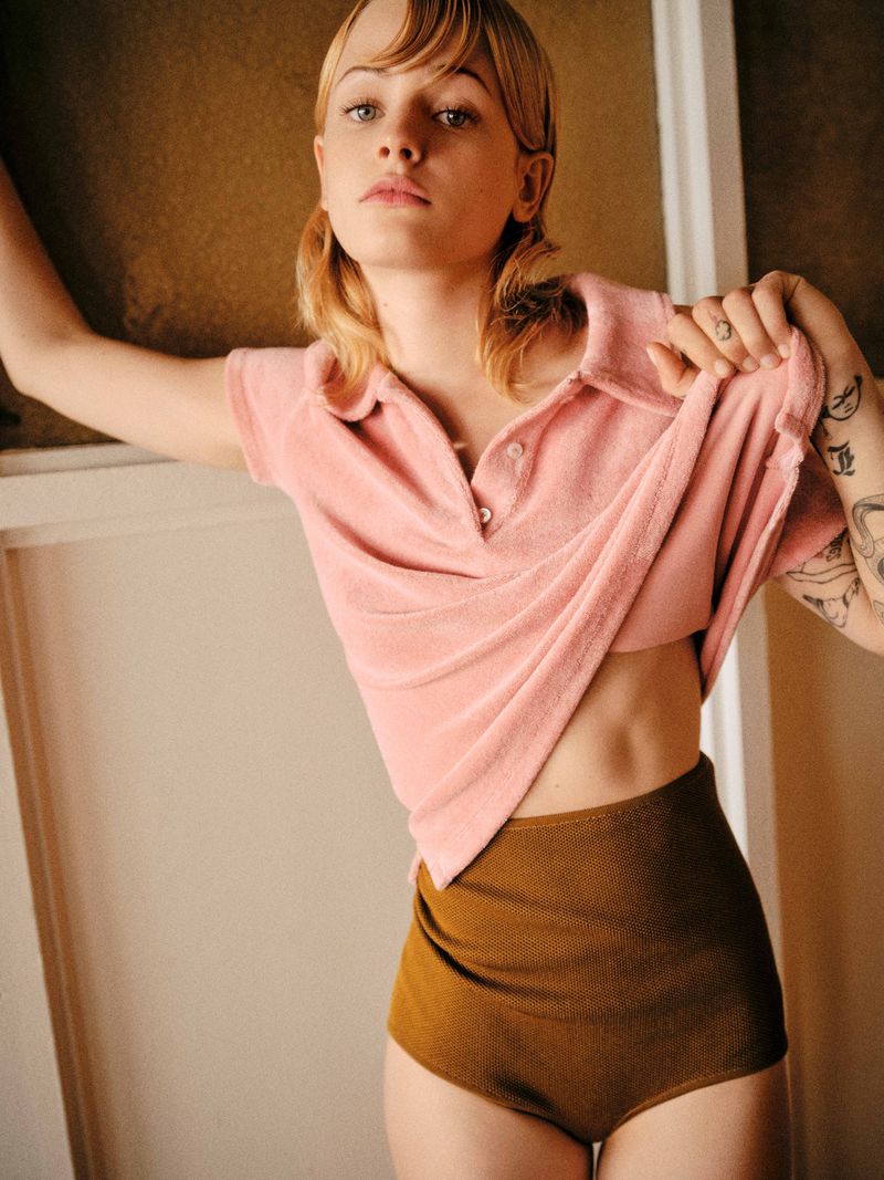 A woman in a pink short-sleeve polo shirt and brown bottoms strikes a pose in an indoor setting, showcasing fashion-forward style.