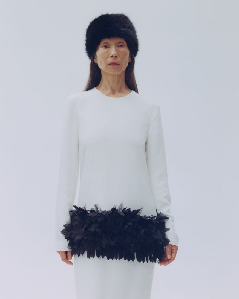 Model wearing a white dress with a black feather trim and black fur hat against a plain white background