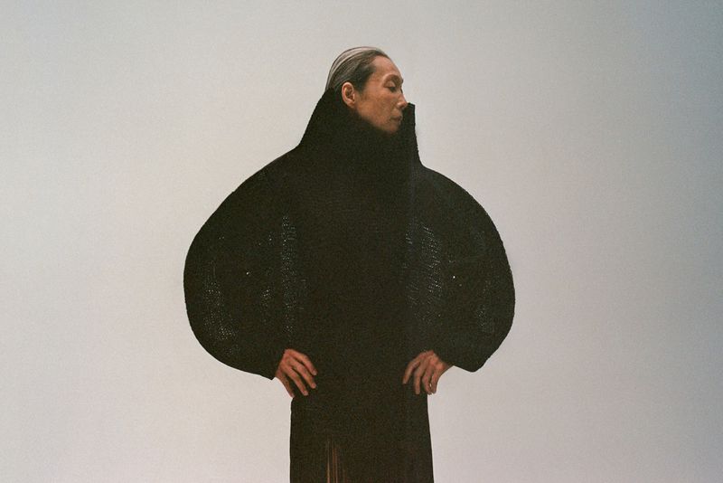 Model wearing a structured black high-collar coat with exaggerated rounded sleeves against a plain grey background