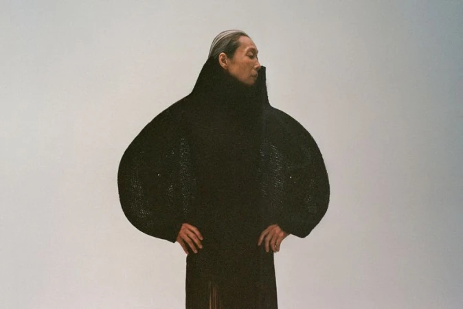 Model wearing a structured black high-collar coat with exaggerated rounded sleeves against a plain grey background