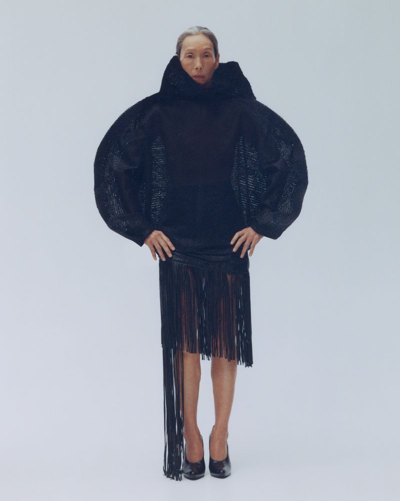 An elderly woman poses in a black, fringed skirt and large-sleeved top against a white background, creating a striking fashion portrait.