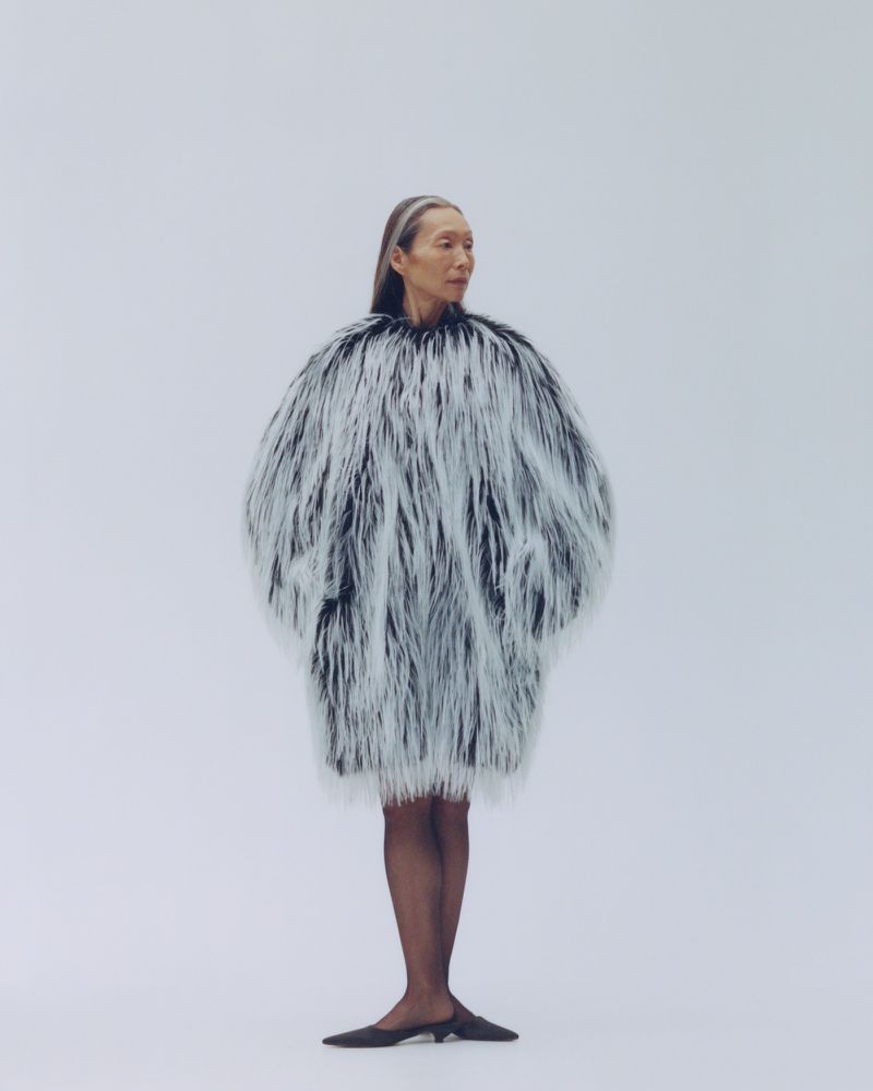 Model wearing a voluminous black and white textured faux fur coat against a plain light grey studio background.