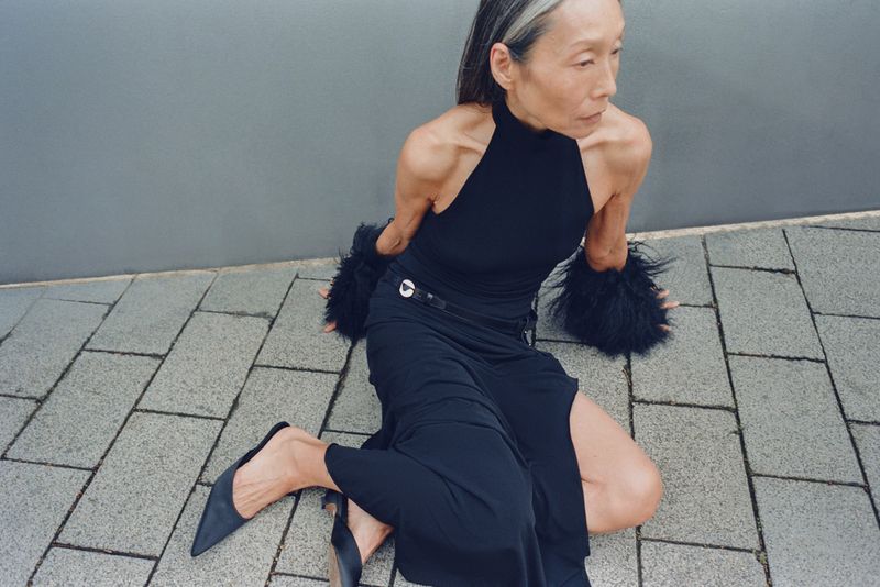 Model Yuri Fukuhara seated on grey stone paving in a black halter top and skirt with furry arm cuffs for Puss Puss Magazine