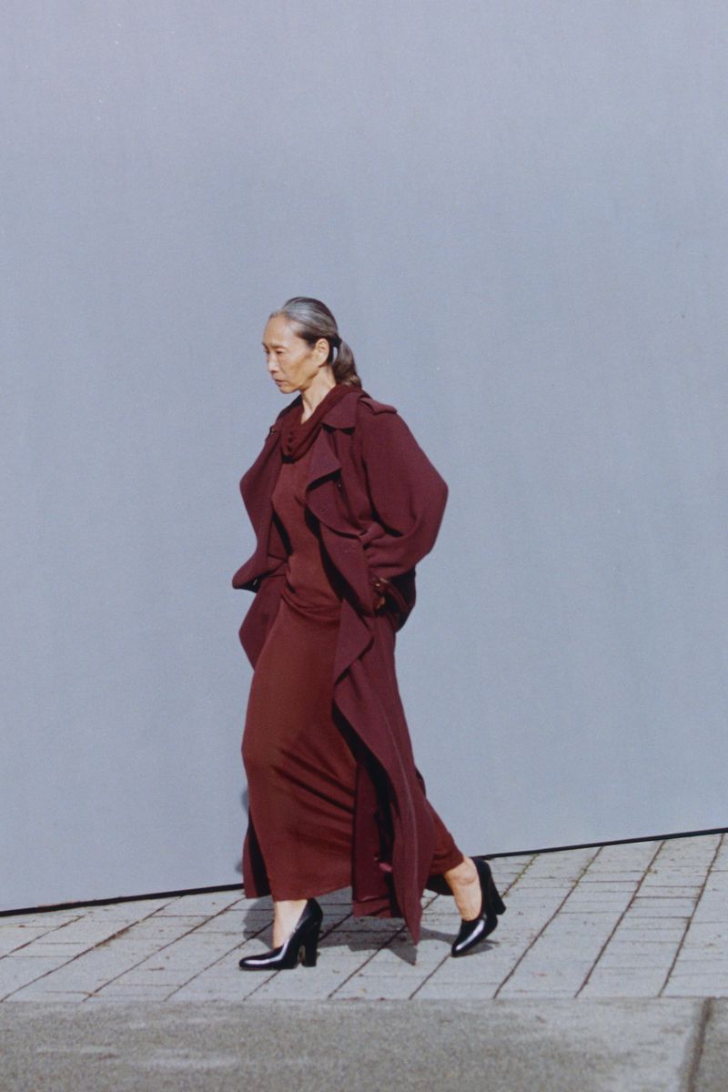 Model Yuri Fukuhara walking in a long burgundy coat against a grey wall for Puss Puss Magazine editorial.