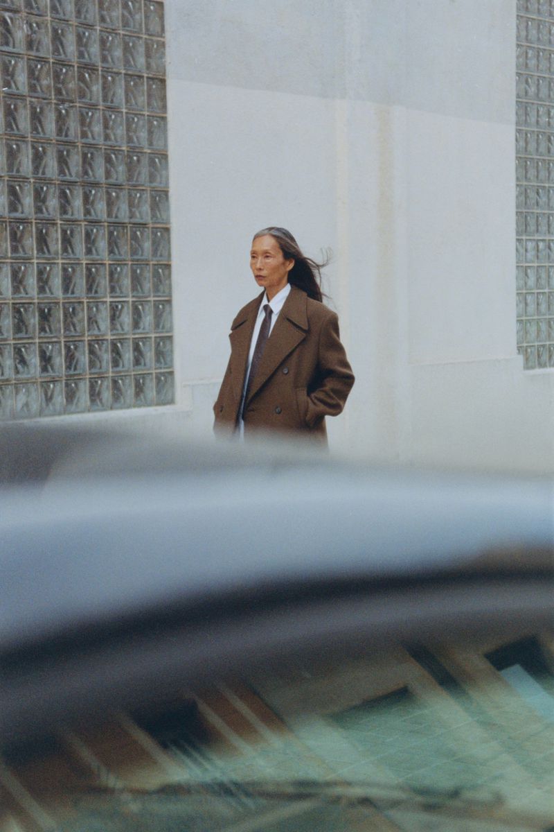 Model in a brown coat and tie walking past a building with glass block windows