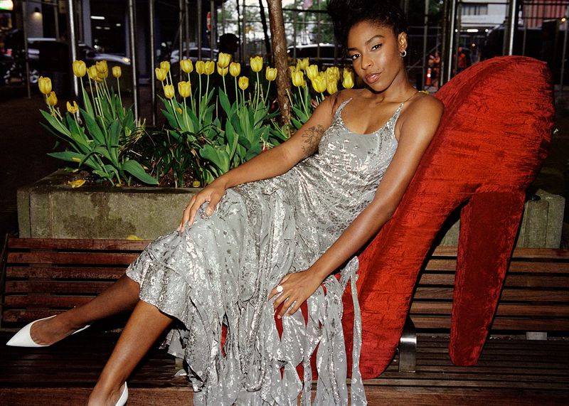 A fashionable Black woman poses on a bench next to a giant red high heel and blooming yellow tulips in a city park at night.