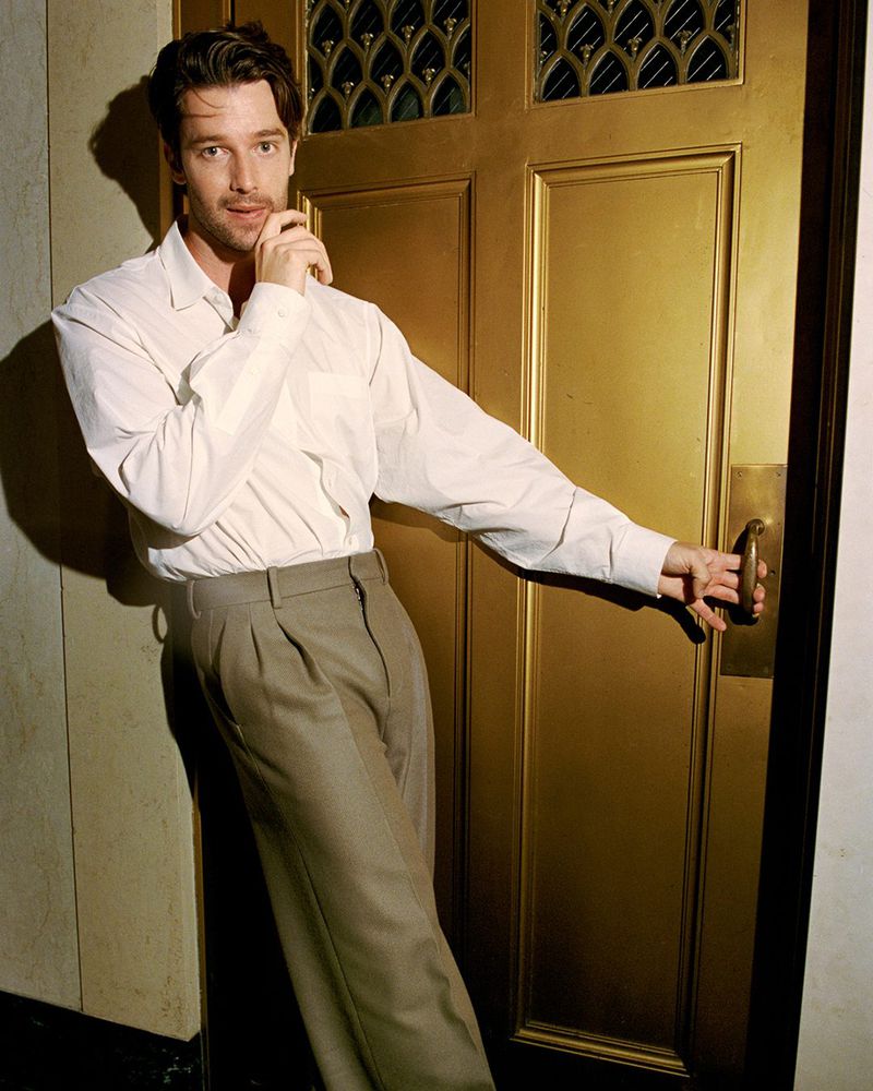 Adult man in white shirt and beige pants leans on golden door, holding the door handle, indoor setting with diffused lighting.
