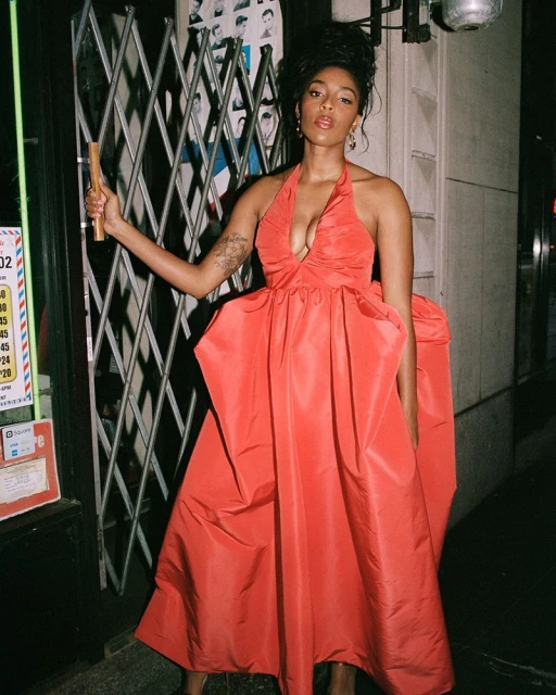 Woman in a coral gown stands outside a building with a barred window at night, holding a wooden object and wearing earrings.