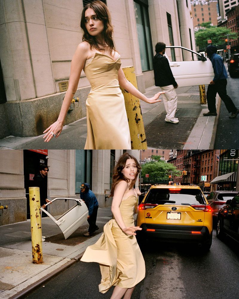A young woman in a champagne-colored gown stands on a city street with yellow taxi and people carrying a car door in the background.