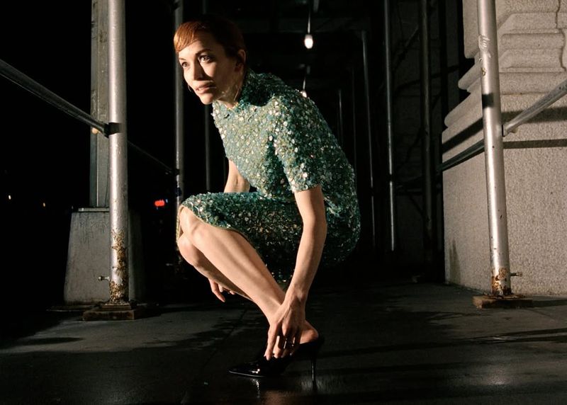 A woman in a sequined dress kneels on a dark street, adjusting her shoe in an outdoor setting with an urban backdrop.