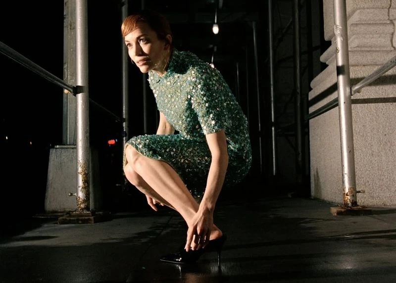 A woman in a sequined dress kneels on a dark street, adjusting her shoe in an outdoor setting with an urban backdrop.